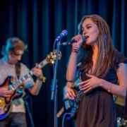 Ein Foto: Eine Bühnensituation. Im Vordergrund eine junge Sängerin, die in ein Mikro singt. Im Hintergrund etwas verschwommen zwei Menschen, die E-Gitarre spielen.