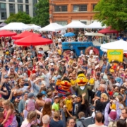Foto: Eine Freifläche umgeben von Backsteinbauten. Es wimmelt vor Menschen. Im Hintergrund sieht man viele, bunte Sonnenschirme. In der Menge sieht man Ernie und Bert aus der Sesamstraße als menschengroße Stoffpuppen.Ein open air Familienfest