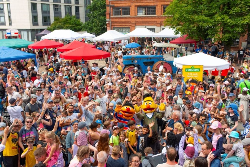 Foto: Eine Freifläche umgeben von Backsteinbauten. Es wimmelt vor Menschen. Im Hintergrund sieht man viele, bunte Sonnenschirme. In der Menge sieht man Ernie und Bert aus der Sesamstraße als menschengroße Stoffpuppen.Ein open air Familienfest