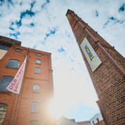 Ein Foto: >Von unten nach oben aufgenommen, Blick in den Himmel, rechts die Fassade vom Museum der Arbeit in Hamburg, links der historiche Schornstein der Zinnschmelze mit einem Banner auf dem TINCON steht.