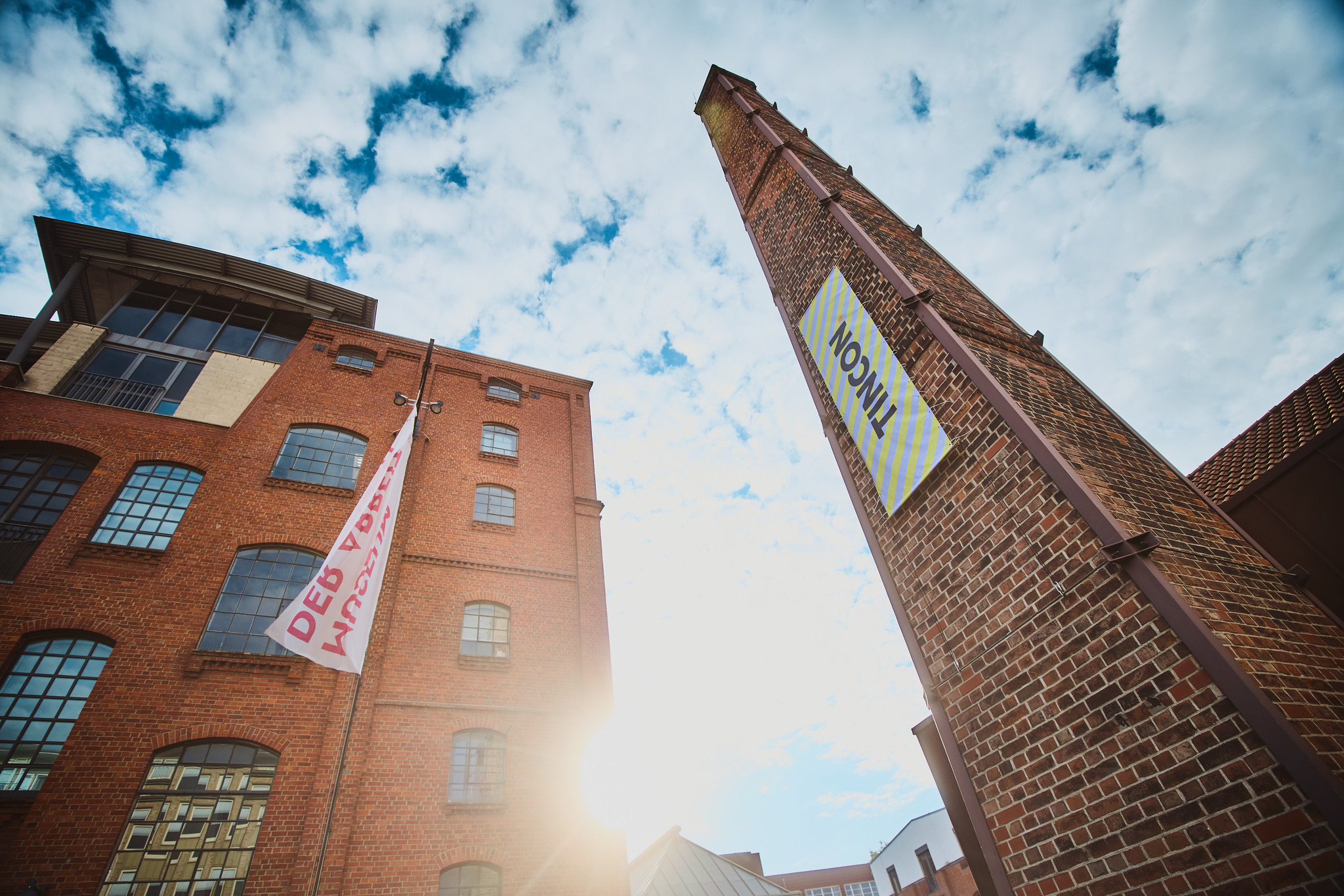 Ein Foto: >Von unten nach oben aufgenommen, Blick in den Himmel, rechts die Fassade vom Museum der Arbeit in Hamburg, links der historiche Schornstein der Zinnschmelze mit einem Banner auf dem TINCON steht.